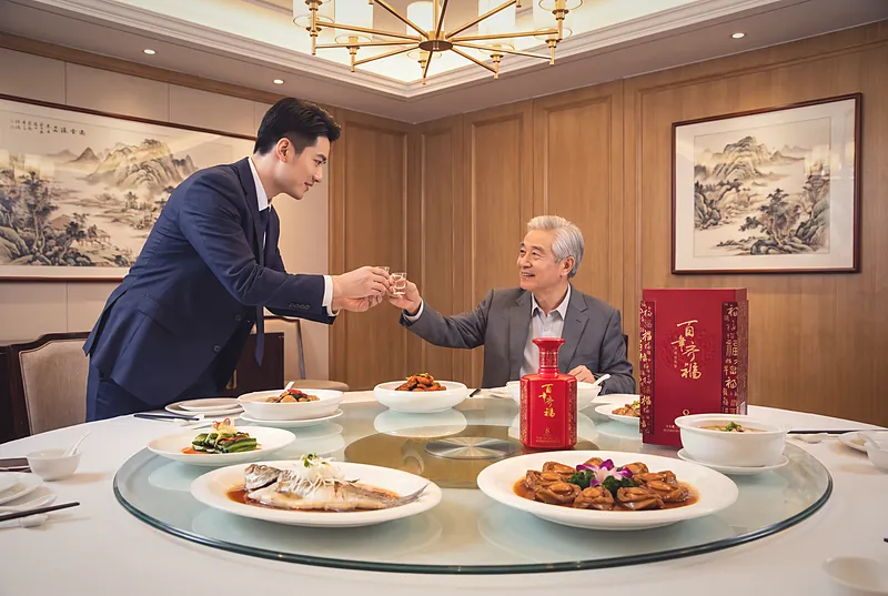 正式的中式商务宴席上，一位宾客在圆桌旁双手举杯向长者敬酒，桌上摆着百年齐福·8